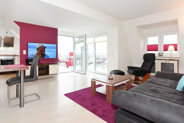 Un salon moderne comprenant un téléviseur, un canapé, un coin repas et un balcon avec des stores rouges.