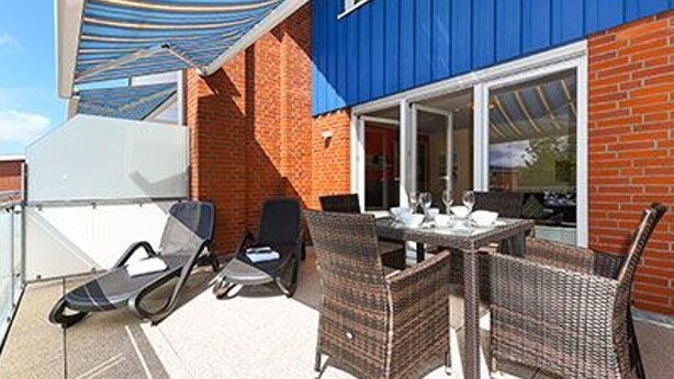 Une terrasse extérieure aménagée avec des meubles en osier, une table dressée pour le repas et un auvent bleu offrant de l’ombre.