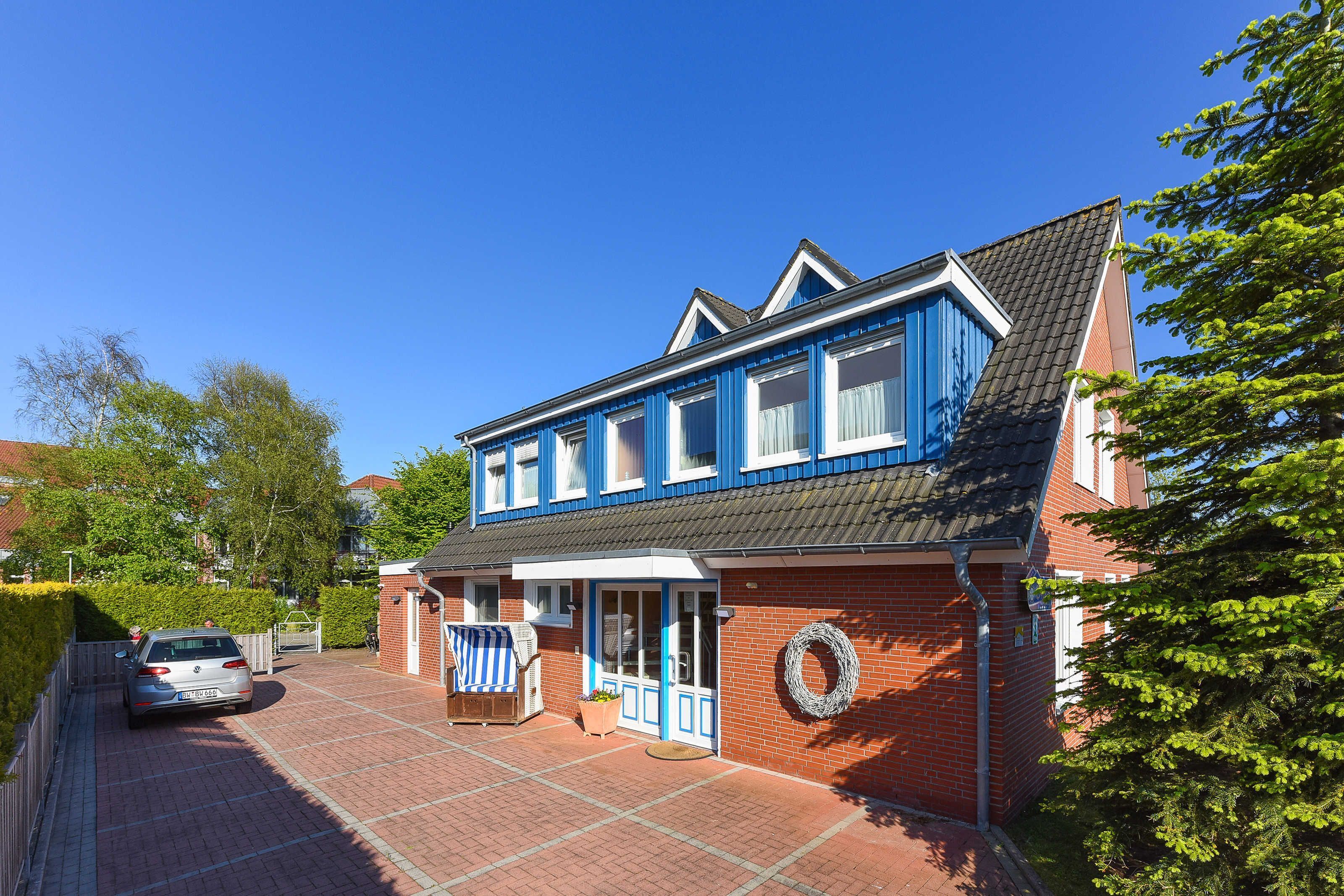 Une maison à deux étages, à la façade bleue et blanche, avec une allée pavée et une voiture garée dans l’allée.