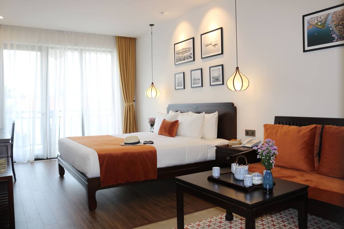 A hotel room with a bed, a sofa, a coffee table, and a desk. There are framed pictures on the wall and a window with curtains.
