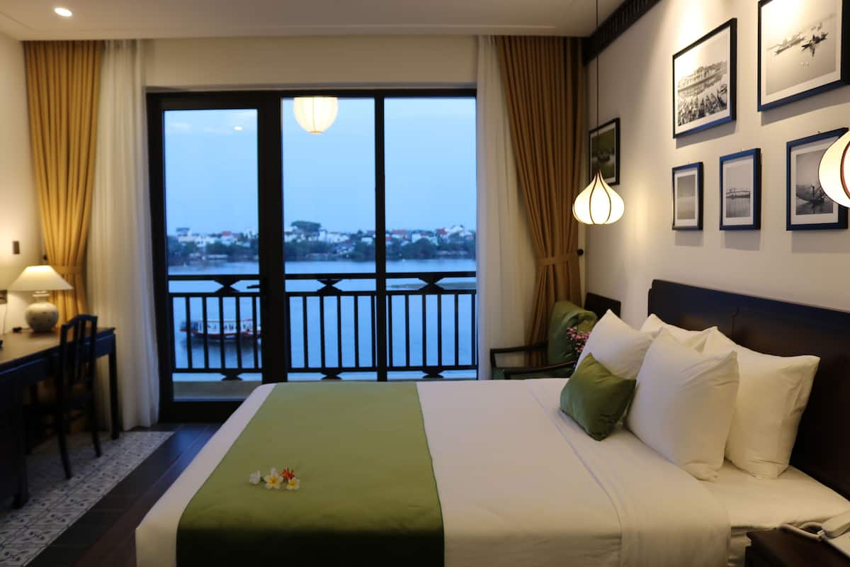 A hotel room with a large bed, a desk, a chair, a view of a body of water through a balcony, and several framed pictures on the wall.