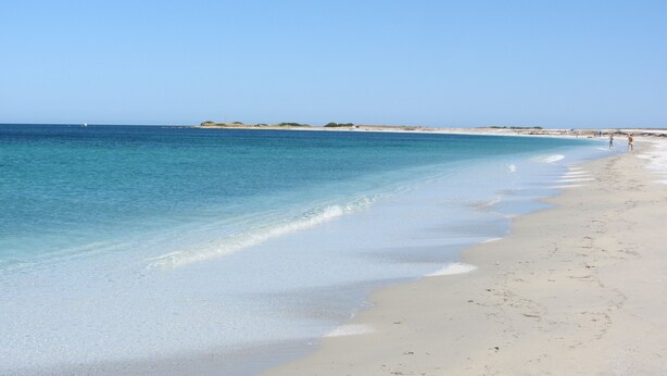 A beach with clear blue water, sandy shore, and a few people walking.