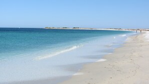 Een strand met helderblauw water, een zandstrand en enkele mensen die wandelen.