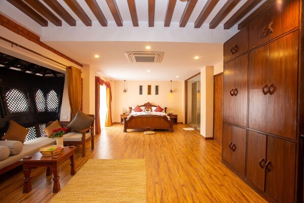 A spacious bedroom with a wooden floor, a large wardrobe, a bed with red bedding, a sitting area with a sofa and armchairs, and a small table with a basket of fruit.