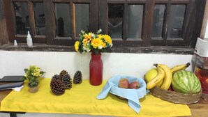 A table with a yellow cloth, a red vase with yellow flowers, pinecones, a blue cloth with fruits, and a basket with bananas and a watermelon.