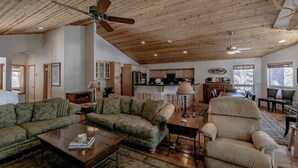 Un salón espacioso con techo alto, ventilador de techo, chimenea y una cocina visible a través de una puerta abierta.