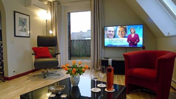 A living room with a TV, a red chair, a black chair with a red pillow, a vase with flowers, and a table with wine glasses and a bottle. - FeWo-Schuberski (Oerlinghausen)