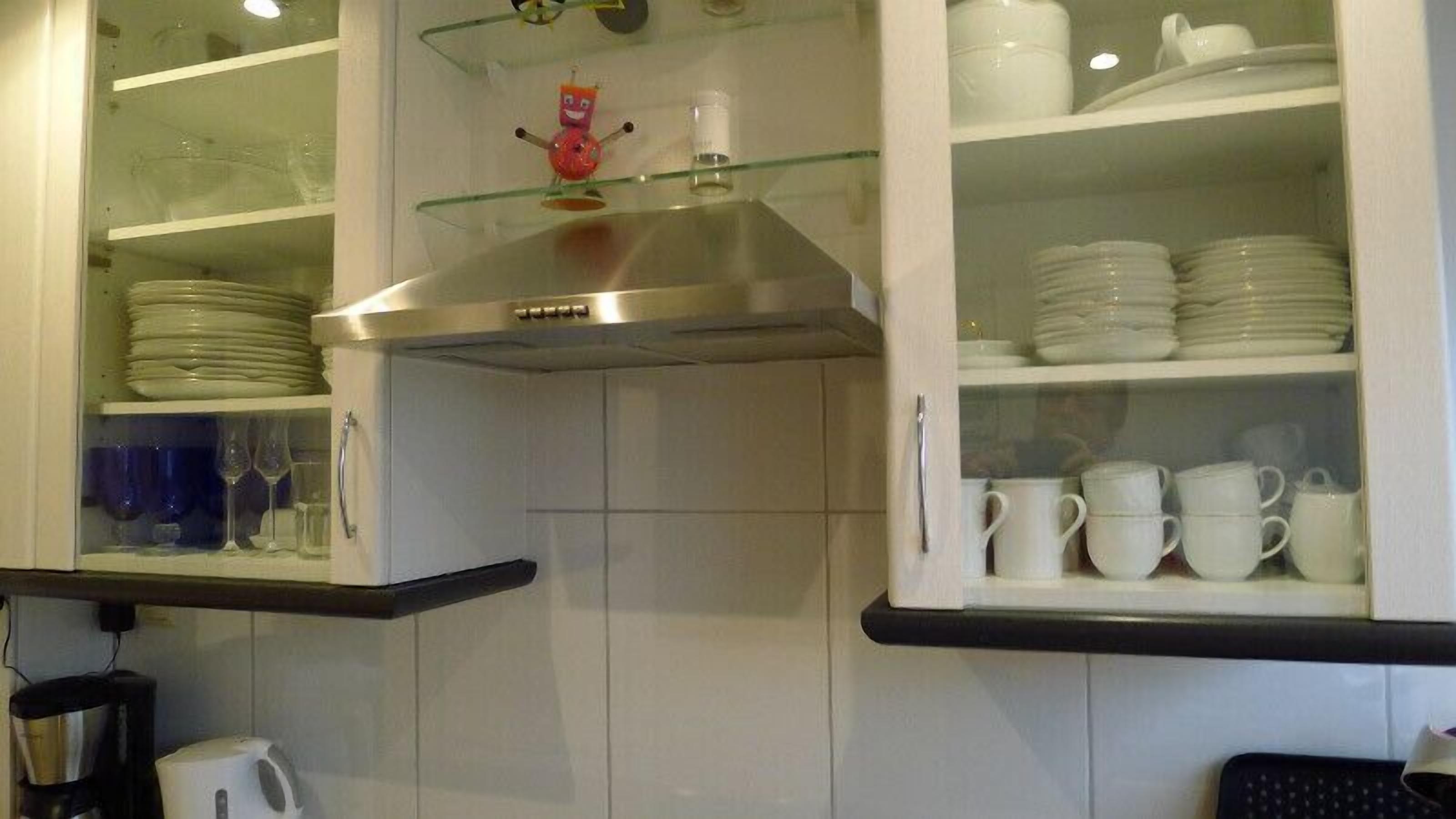 A kitchen with white cabinets, a stainless steel range hood, and glass shelves holding plates, cups, and a small toy.