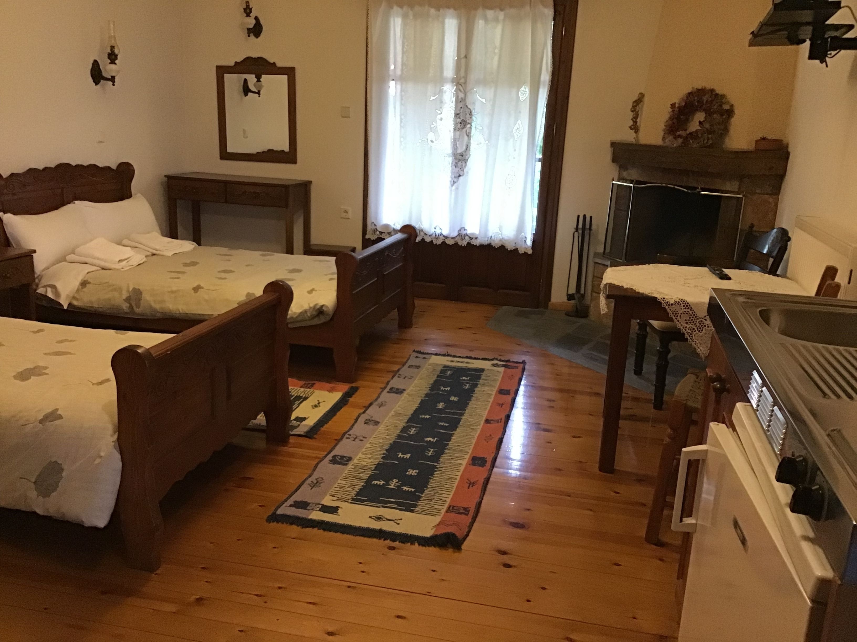 A room with two wooden beds, a small table, a fireplace, and a kitchen area.