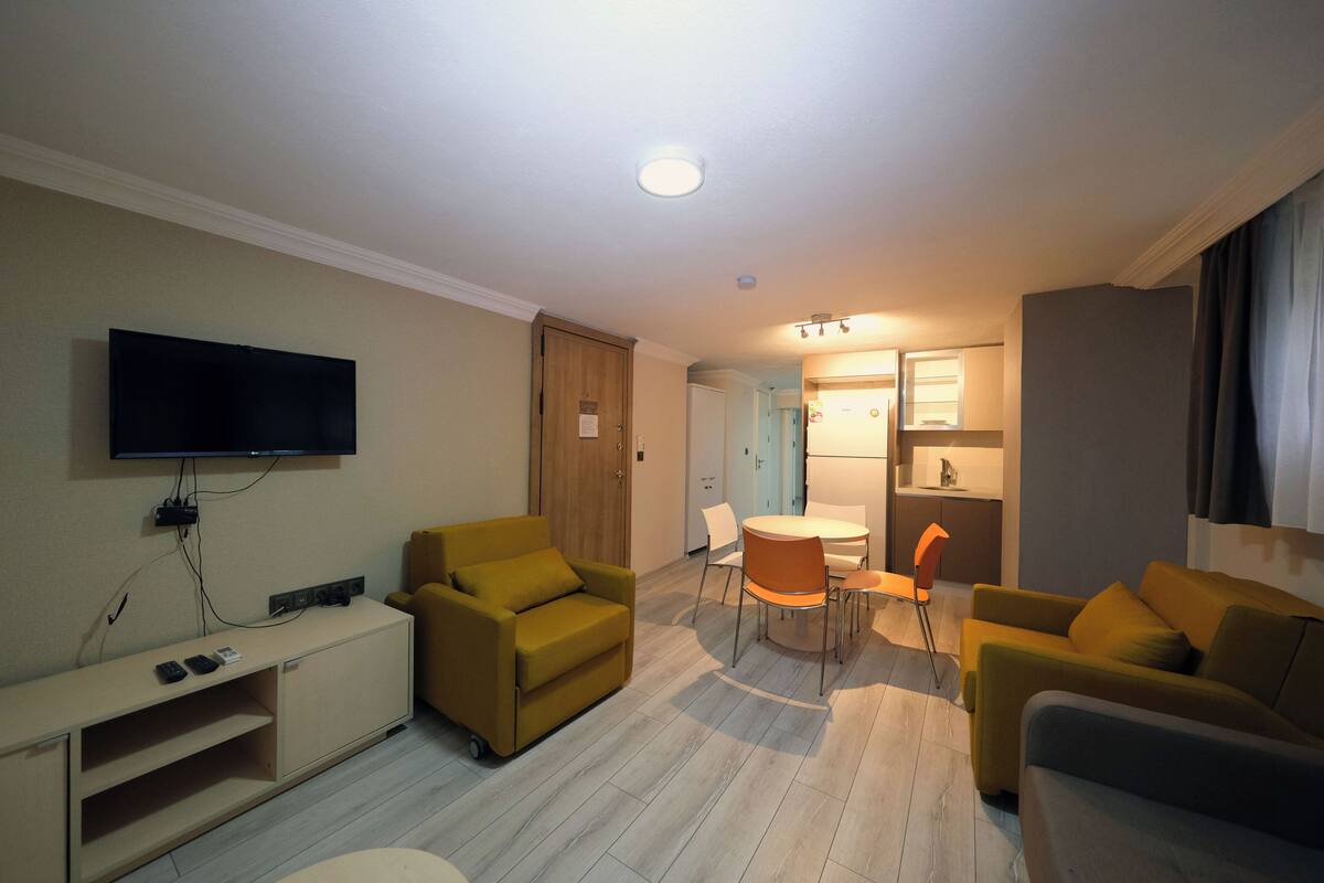 Un salon moderne avec un téléviseur à écran plat, un canapé, une table à manger avec des chaises, et un coin cuisine à l’arrière-plan.