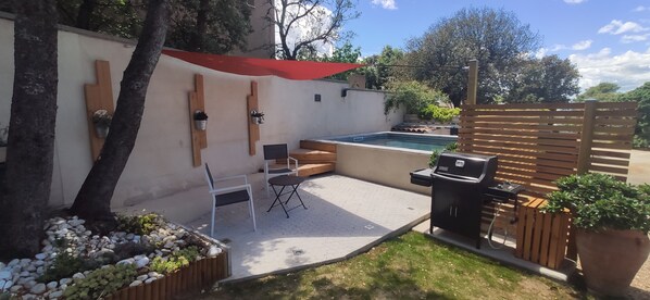 Un jardin avec une piscine, un barbecue, une terrasse en bois et un auvent rouge.