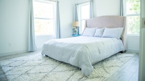 Ein Schlafzimmer mit einem großen Bett, einem Nachttisch mit Lampe und zwei mit Vorhängen versehenen Fenstern.