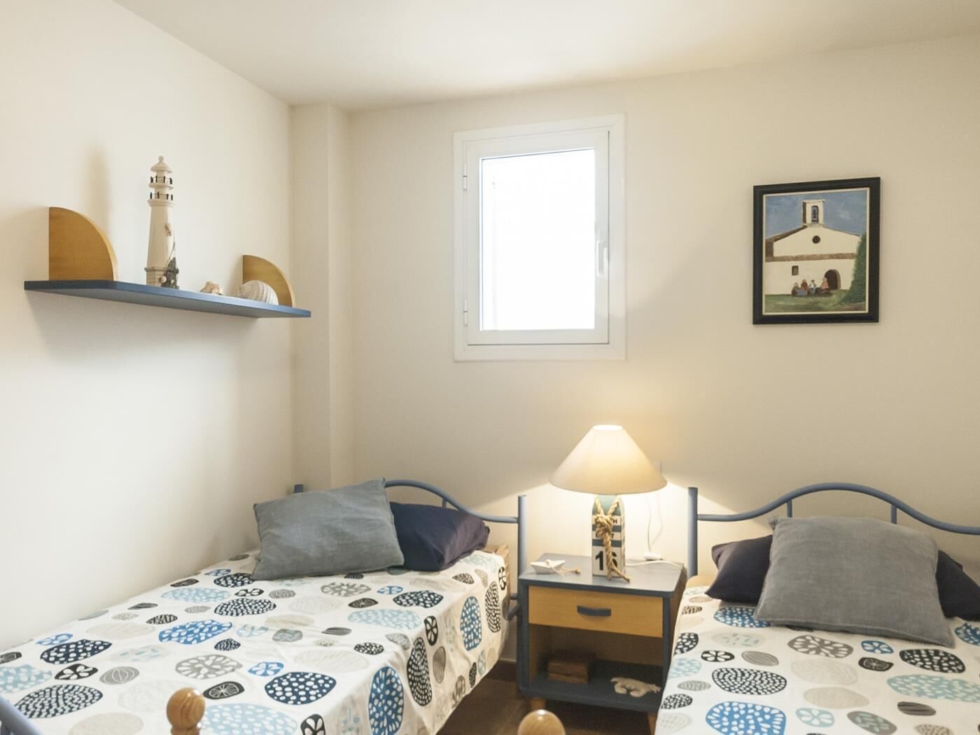 A room with two single beds, a nightstand with a lamp, a window, and a wall-mounted shelf with decorative items.