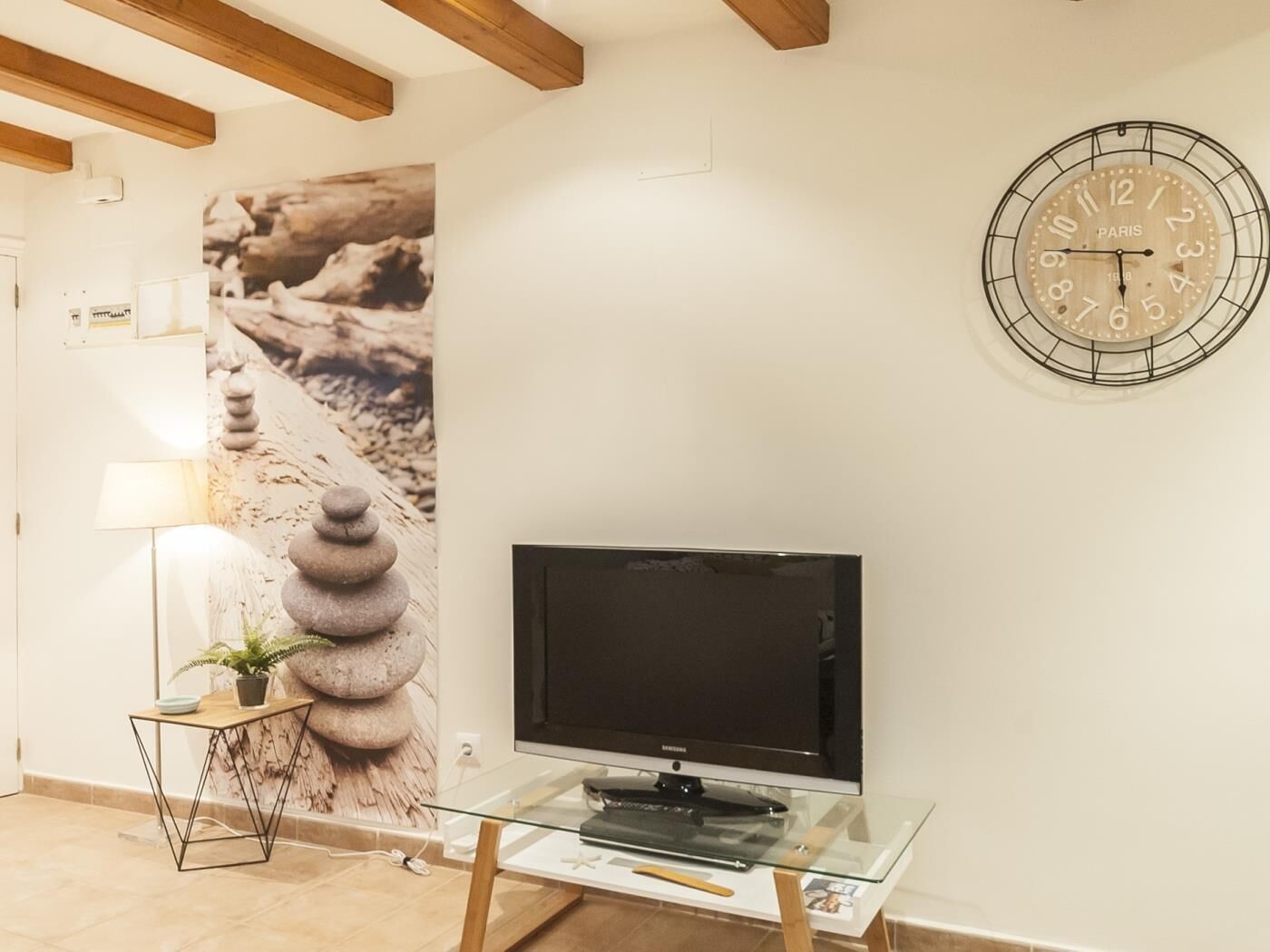 A modern living room with a flat-screen TV on a glass table, a wall clock, and a framed picture.