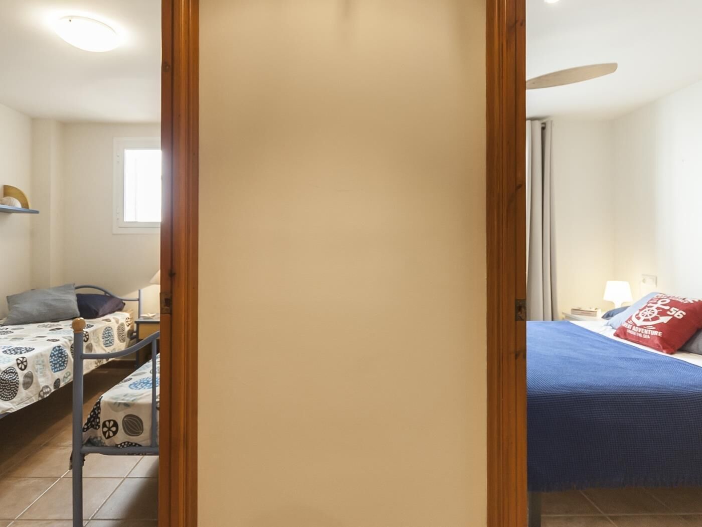 A room with two beds, one with a blue bedspread and red pillows, and another with a patterned bedspread. There is a wooden door in the foreground, and a small table with a chair in the background.