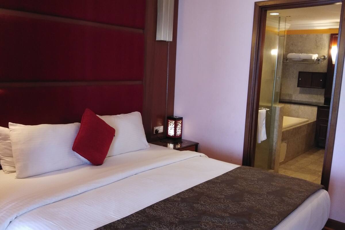 Ein Hotelzimmer mit einem großen Bett, einem roten Zierkissen, einem Nachttisch mit Lampe und Blick ins Badezimmer durch eine Glastür.