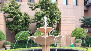 A fountain with water jets in a circular basin, surrounded by greenery and a building with large windows in the background. - Times Square Residence At Kuala Lumpur (Kuala Lumpur)