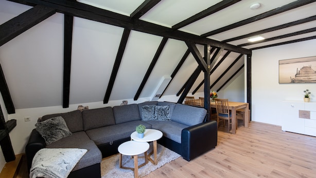 Ein modernes Loft-Schlafzimmer mit freiliegenden Holzbalken, einem grauen Ecksofa, einem kleinen runden Tisch und einem Holzessttisch.