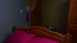 A bedroom with a wooden bed frame, purple bedding, and a nightstand with a bottle. - Kimis Place (Douala)