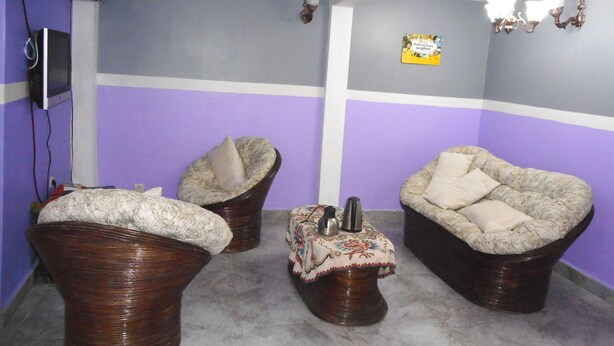 A room with purple walls, two wicker chairs with cushions, a small table with a decorative cloth, and a wall-mounted speaker. - Kimis Place (Douala)