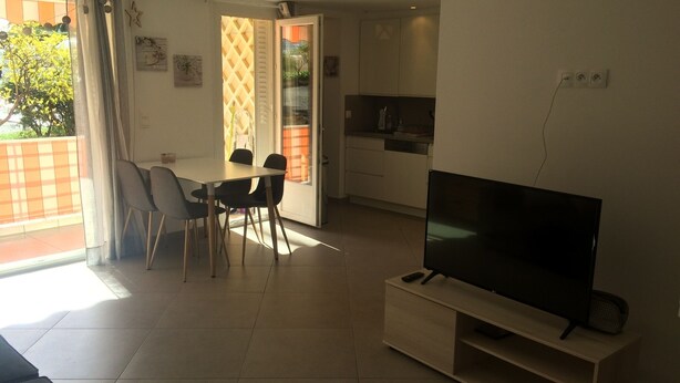 Ein modernes Wohnzimmer mit einem Flachbildfernseher auf einem Standfuß, einem Essbereich mit Tisch und Stühlen sowie einer Küchenzeile mit Schränken und einer Arbeitsplatte.