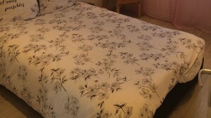 Ein Schlafzimmer mit floralem Bettbezug, einem Korbsessel, einem Wandbild und einem Fenster mit Vorhängen.