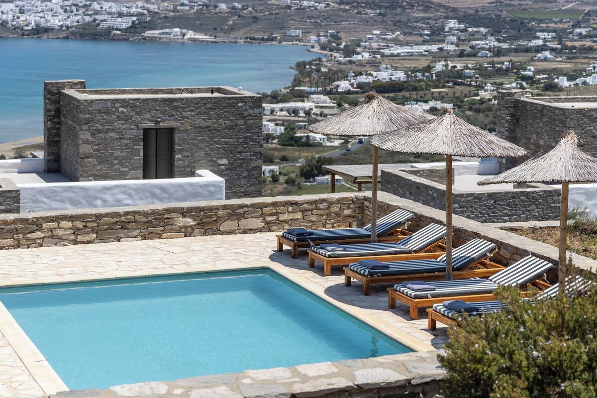 Un espace piscine doté de chaises longues et de parasols, de murs en pierre, et offrant une vue sur la mer et les bâtiments en arrière-plan.