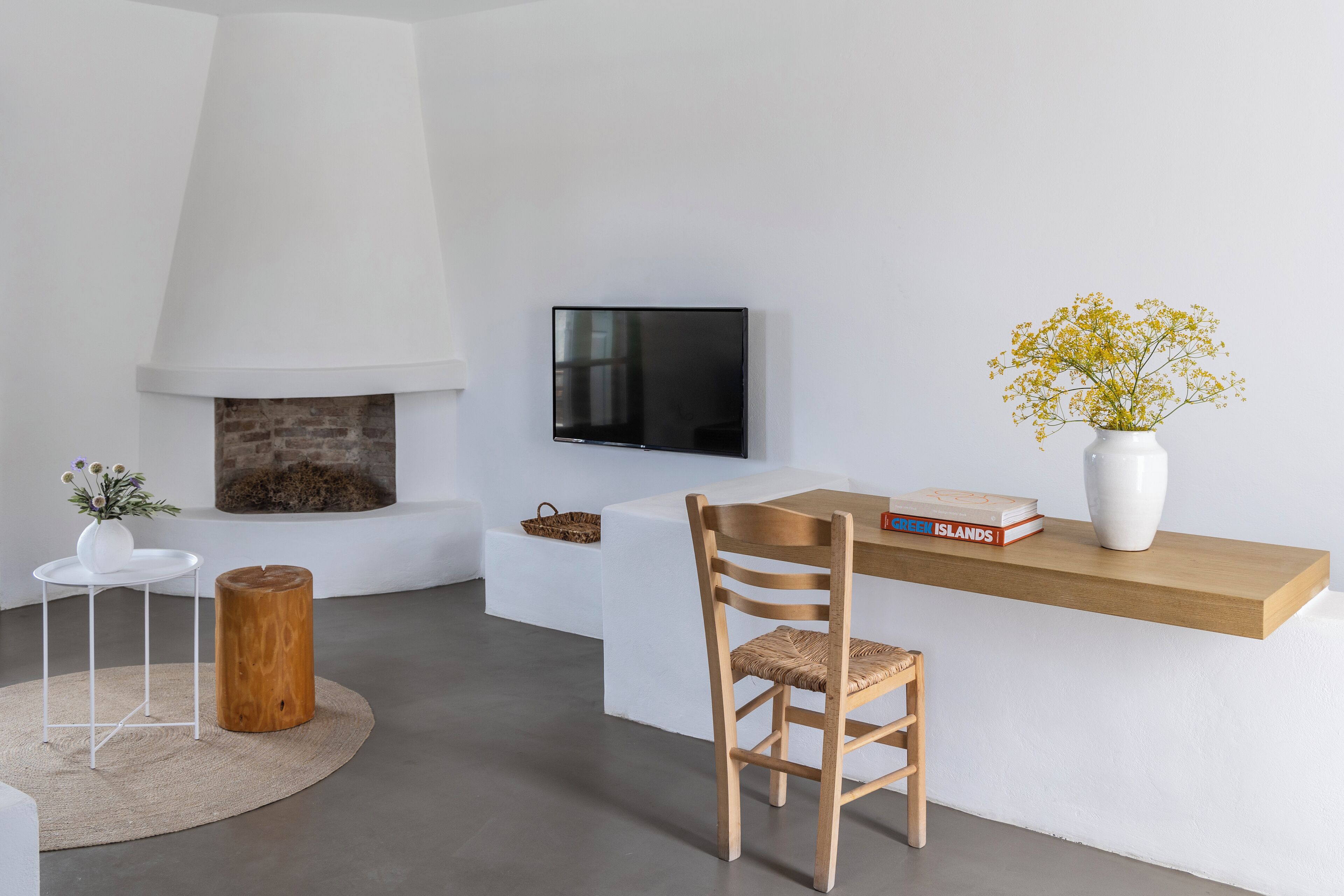 A modern living room with a fireplace, a TV mounted on the wall, a wooden chair, a small round table, and a shelf with books and a vase of flowers.