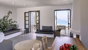 A modern living room with a sofa, a dining table with a plate of tomatoes and peppers, and a view of the sea through open doors.