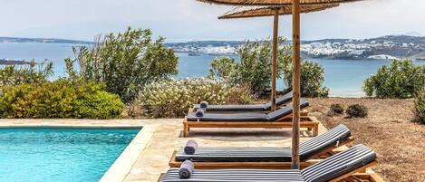 A poolside area with lounge chairs and a thatched umbrella, overlooking a coastal landscape.