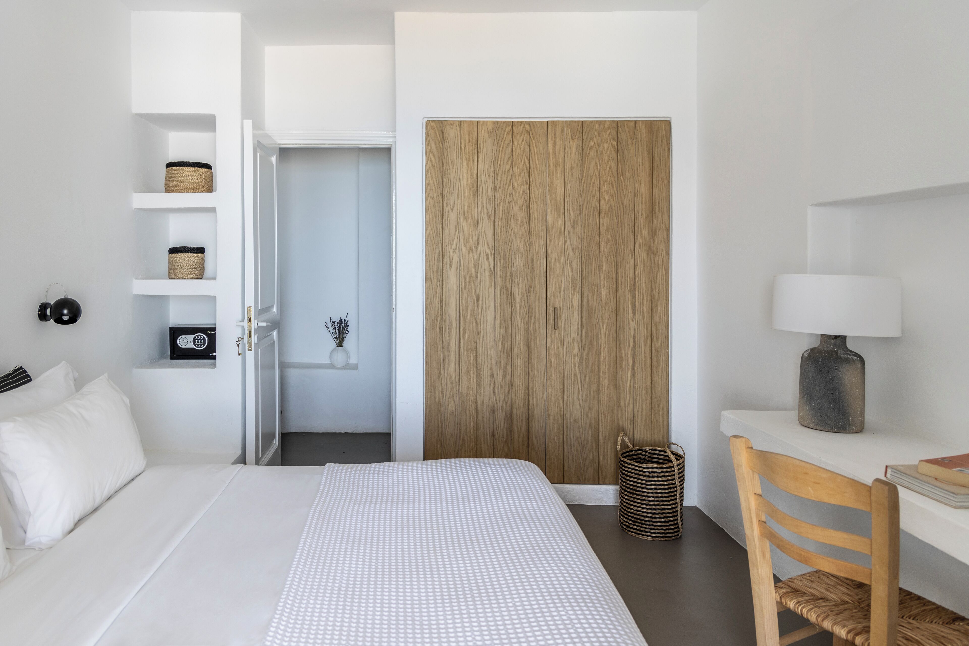 A modern bedroom with a bed, a wooden wardrobe, a desk with a lamp, and shelves with baskets.