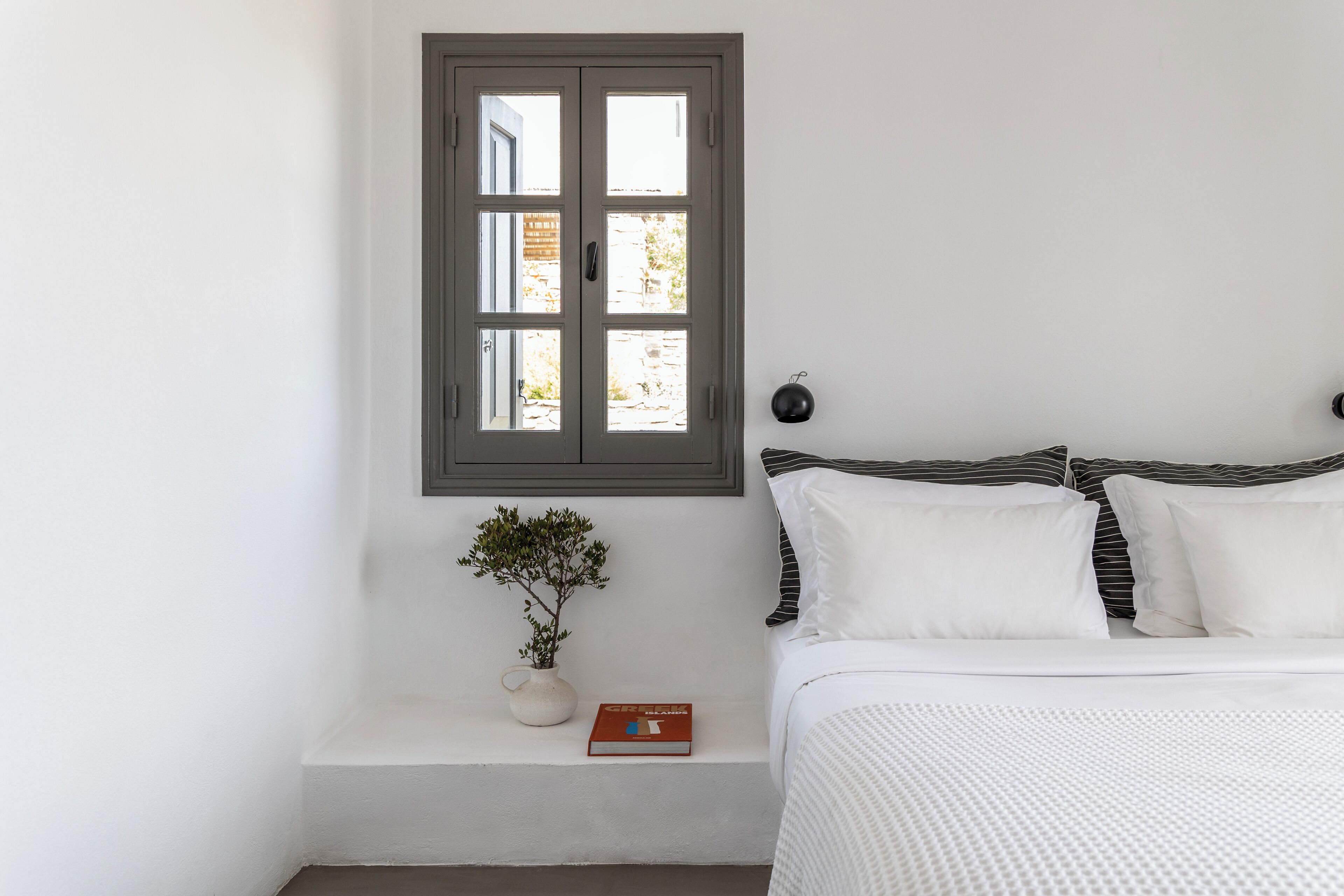 A bedroom with a bed, a bedside table with a book and a plant, a window, and a wall-mounted light.