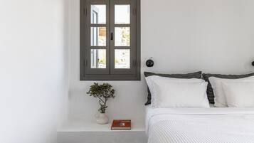 A bedroom with a bed, a bedside table with a book and a plant, a window, and a wall-mounted light.