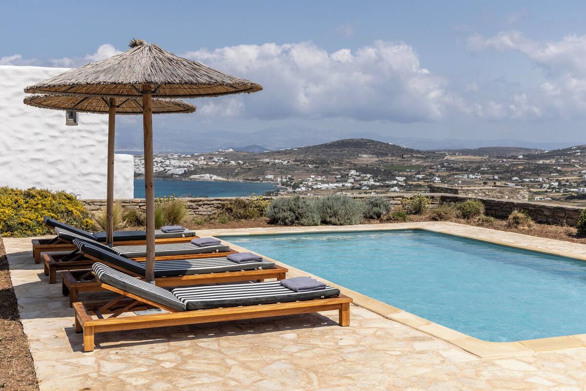Un espace piscine doté de chaises longues et d’un parasol en chaume, offrant une vue sur un paysage côtier.