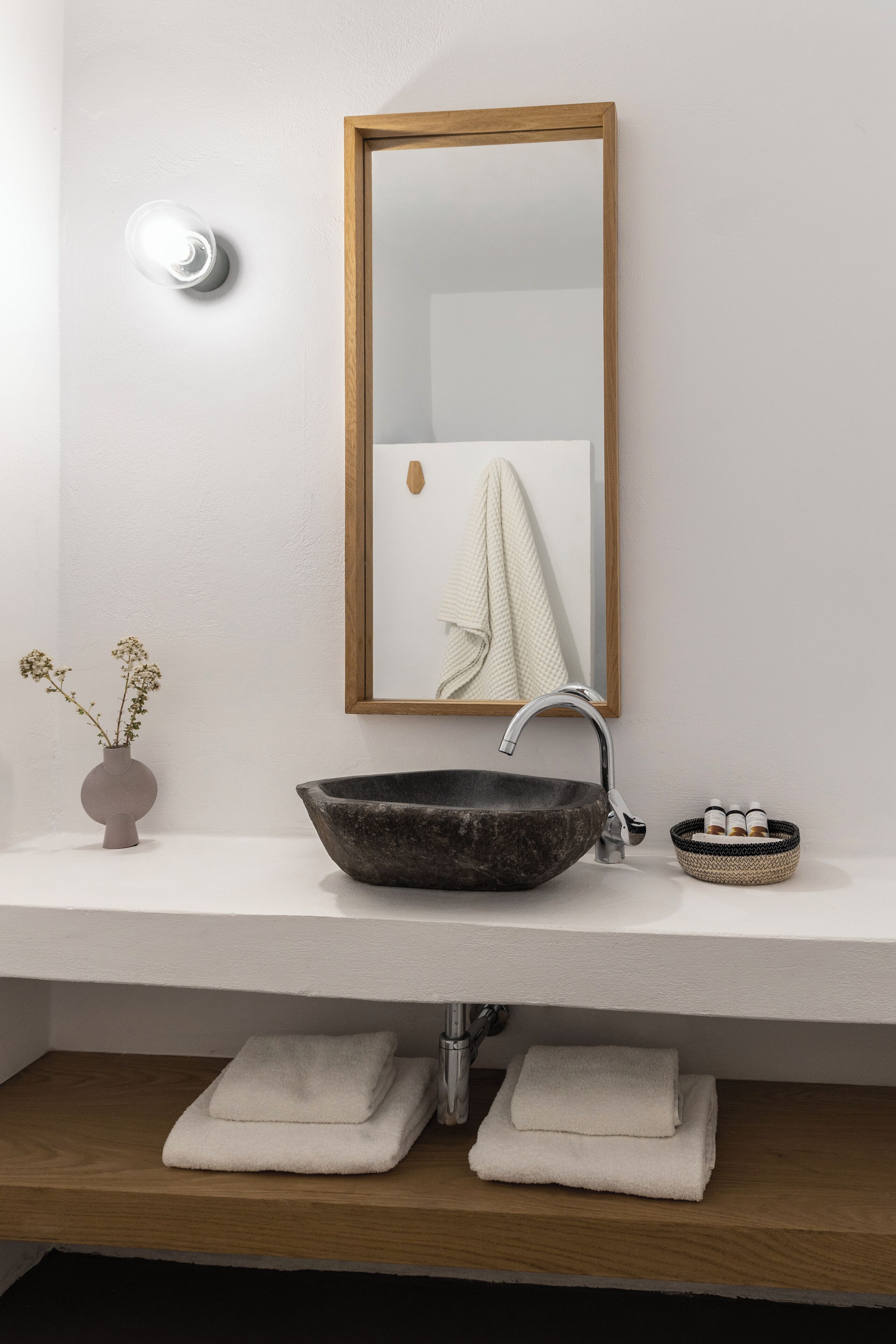 A bathroom with a wooden-framed mirror, a dark stone sink, and a small vase with dried flowers on the countertop.