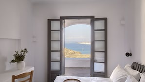 A bedroom with a bed, a chair, a small table with a plant, and a view of the sea through a door with frosted glass panels.