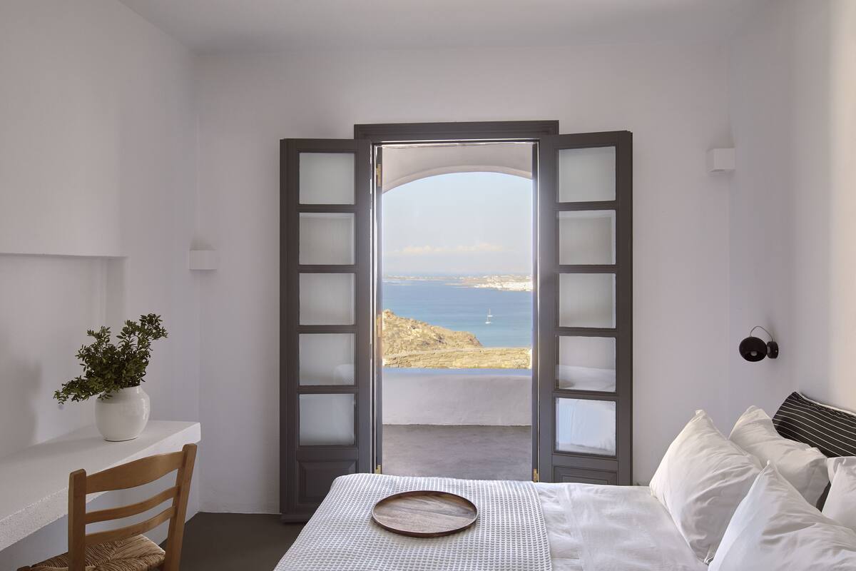 Une chambre à coucher comprenant un lit, une chaise, une petite table avec une plante, et une vue sur la mer par une porte dont les panneaux sont en verre dépoli.