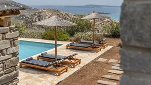 A poolside area with lounge chairs and umbrellas, overlooking a rocky landscape and the sea.