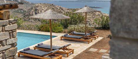 A poolside area with lounge chairs and umbrellas, overlooking a rocky landscape and the sea.