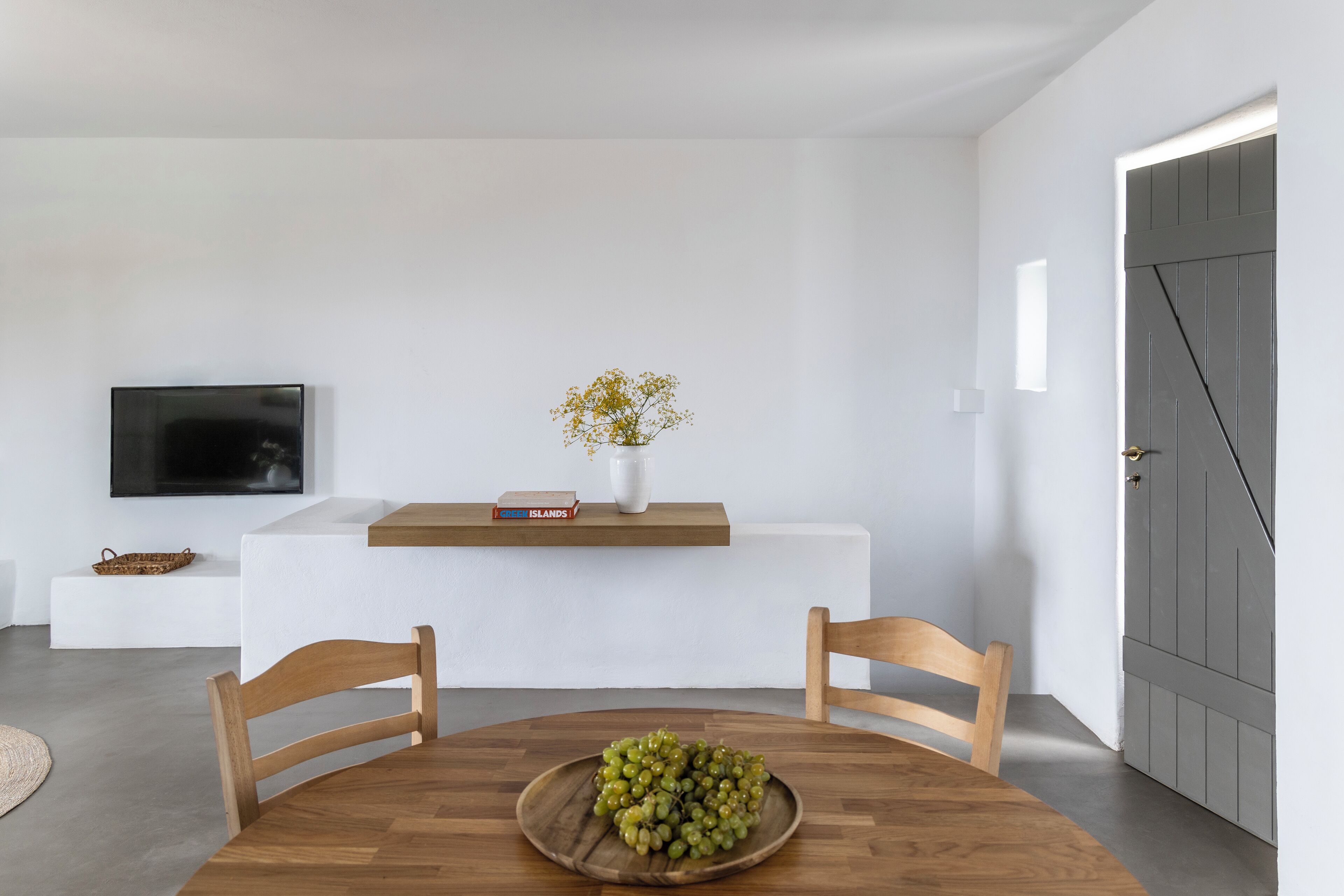 A modern dining area with a wooden table, two chairs, a bowl of green grapes, a white vase with flowers, a flat-screen TV, and a bookshelf with books and a basket.