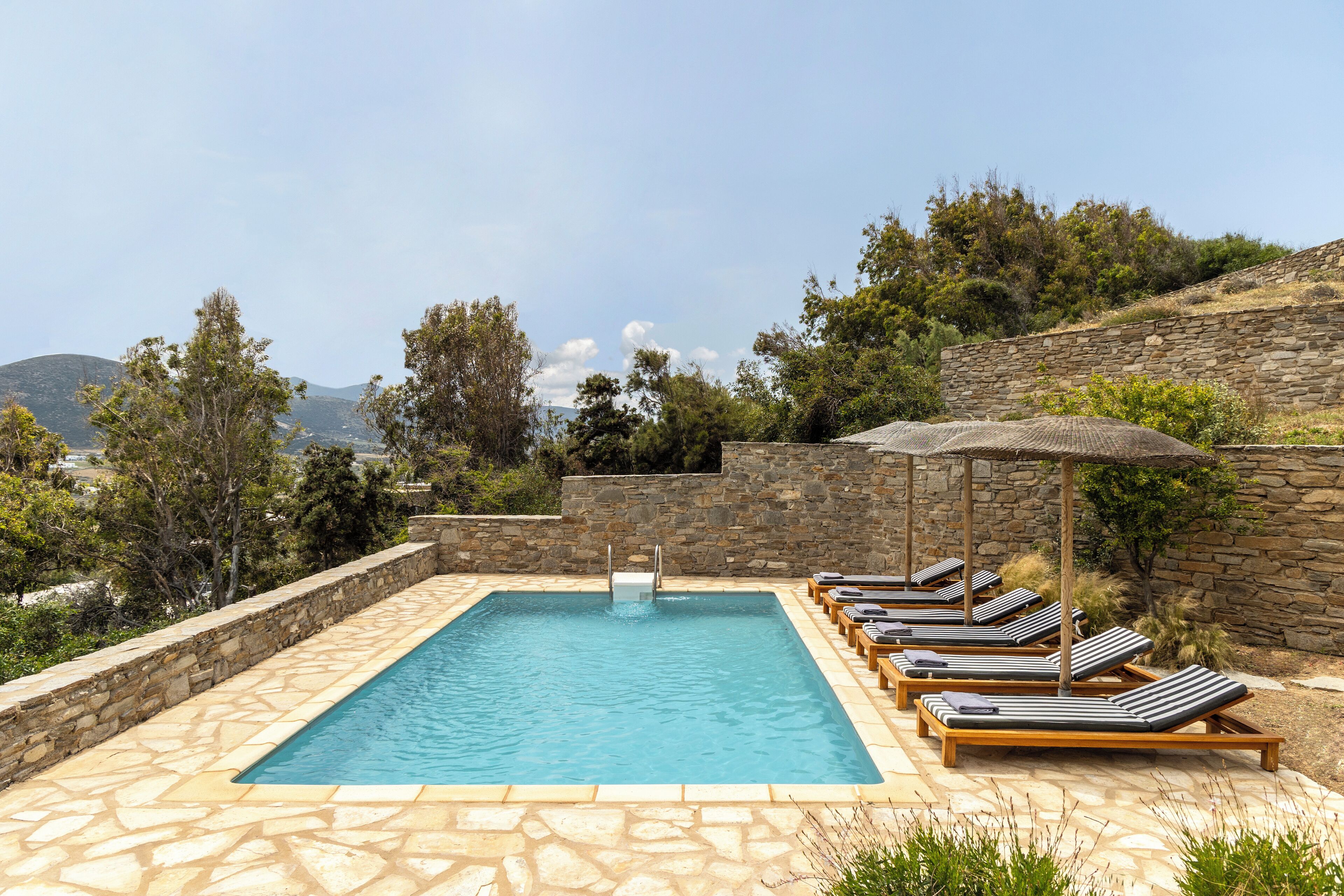 A rectangular outdoor swimming pool with a stone border, surrounded by lounge chairs and umbrellas, set against a backdrop of trees and hills.