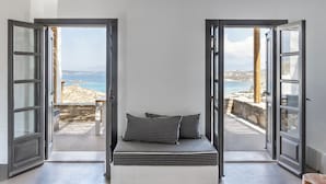 A bedroom with a bed, two open doors leading to a balcony with a view of the sea and a stone wall.