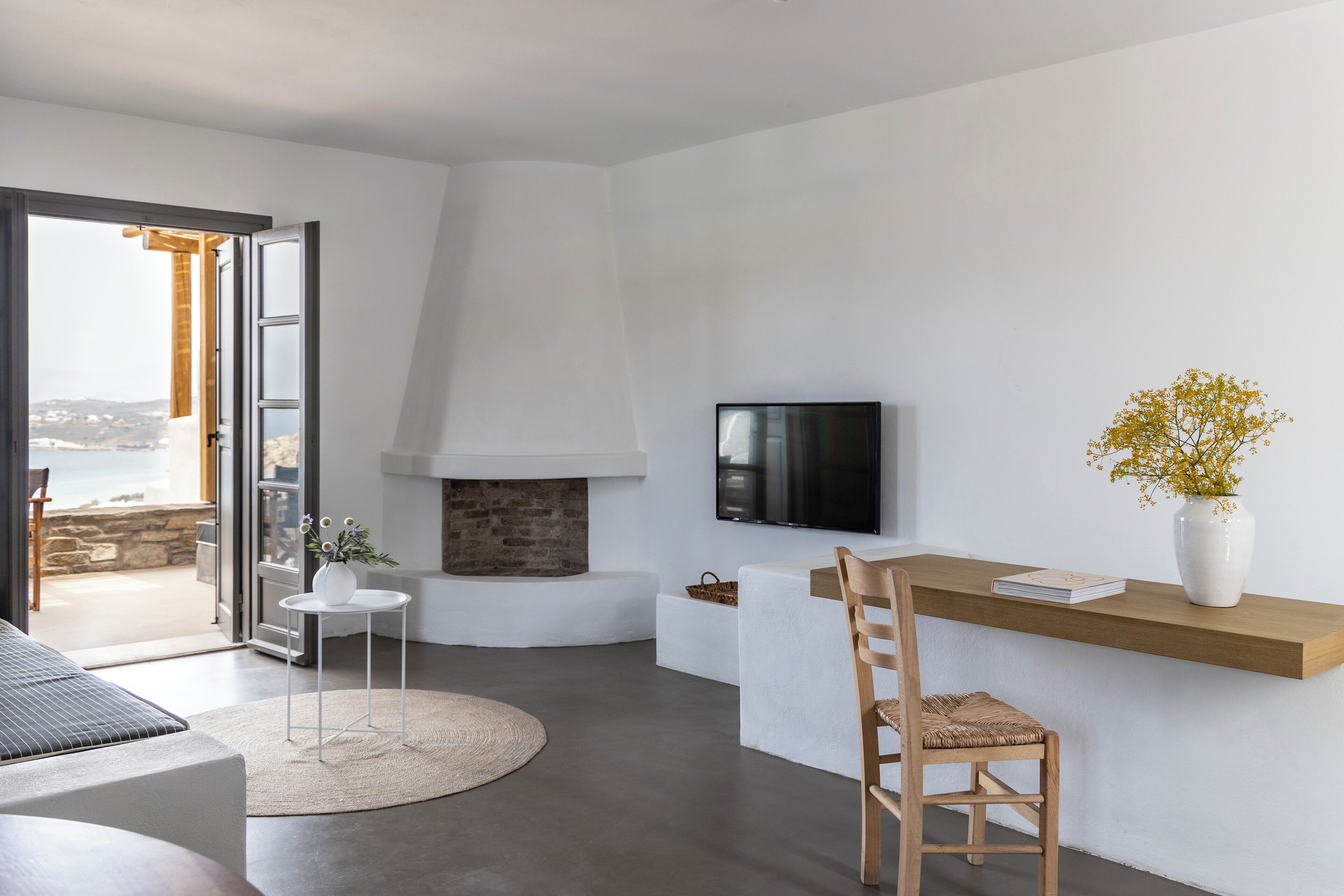 A modern living room with a fireplace, a TV mounted on the wall, a dining area with a wooden table and chairs, and a view of the sea through an open door.
