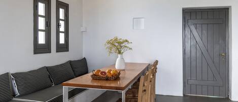A dining area with a long wooden table, a white vase with flowers, and a basket of fruits. There is a dark wooden door and a white wall with two windows.