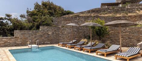 A pool area with lounge chairs and umbrellas, surrounded by stone walls and vegetation.