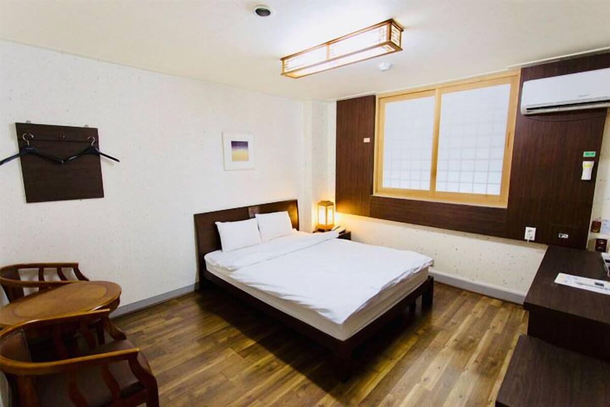 Une chambre d’hôtel avec un lit, un plancher en bois, un bureau, une chaise, une fenêtre et un climatiseur fixé au mur.