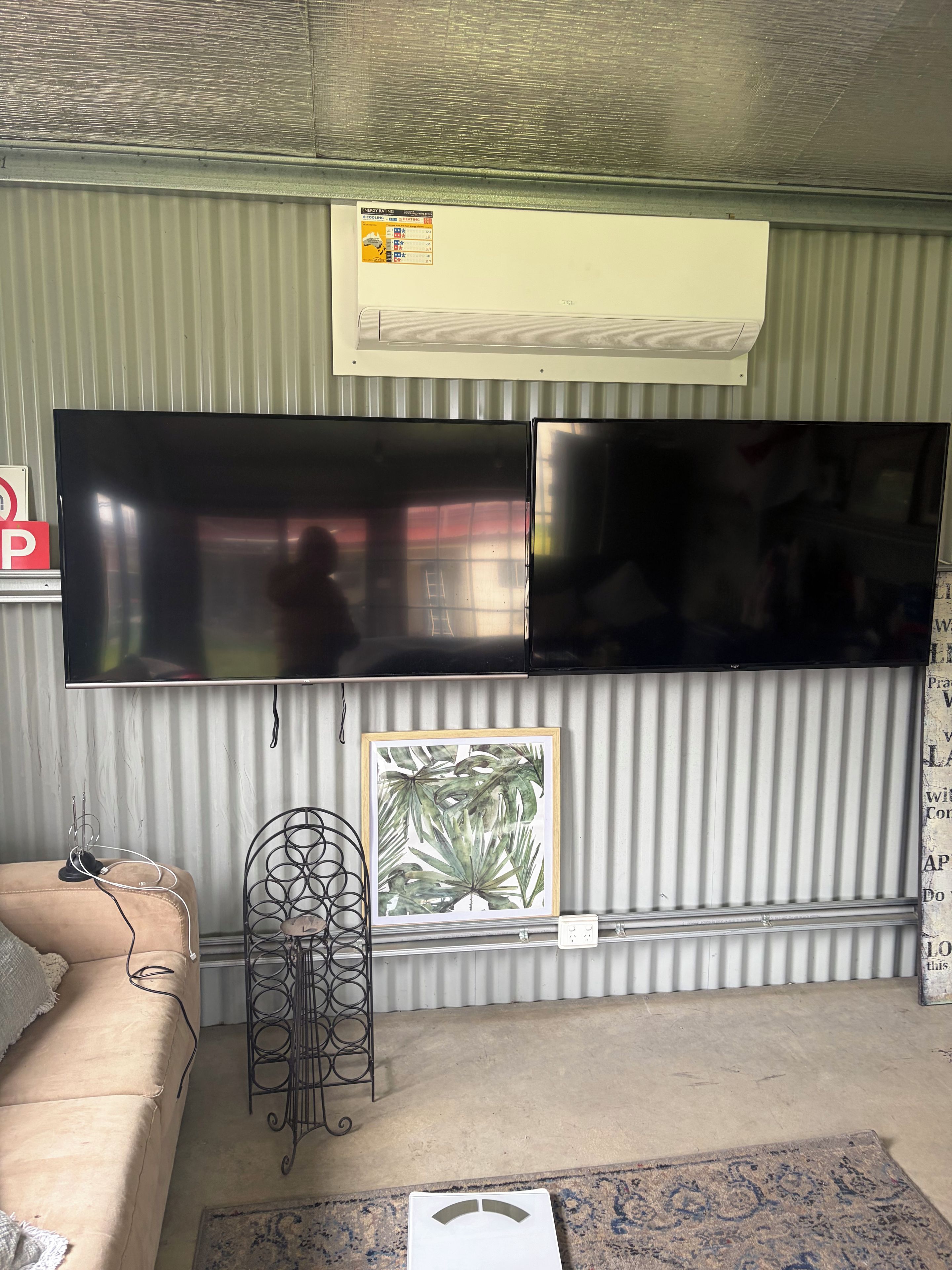 A room with a large flat-screen TV mounted on the wall, a beige sofa, a framed tropical leaf artwork, and a metal stand with a cable.