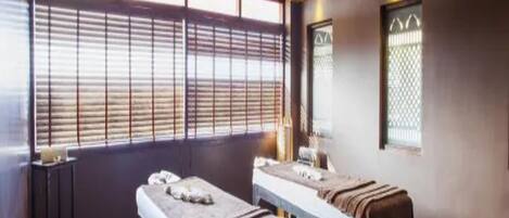 A room with two beds, wooden ceiling, large windows with blinds, and a small table with a lamp.