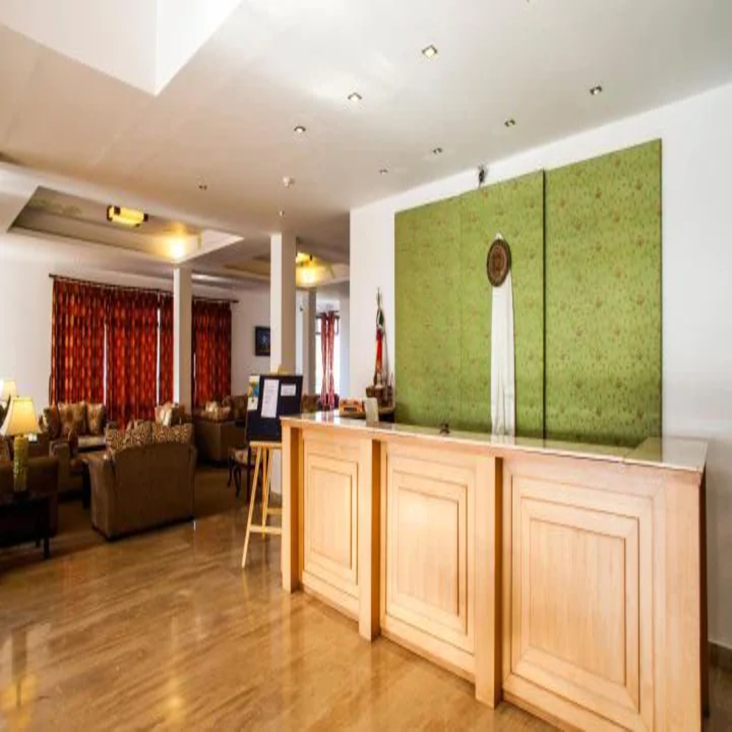 Eine großzügige Hotelhalle mit einem Holzbüro, einer grünen Akzentwand und einer Sitzecke mit Sofa und Vorhängen.