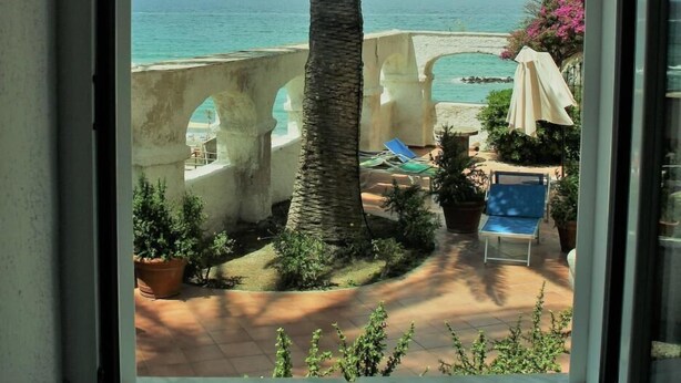 A view from a window overlooking a patio with a palm tree, a stone archway, and a pool area. - Eremo di Montevergine : Calipso, cozy apartment with panoramic view (Forio)
